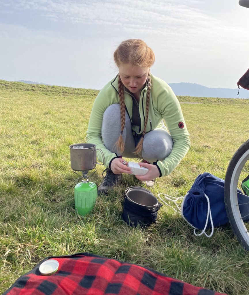 Samantha ist auf einer Wiese zu sehen. Sie ist in der Hocke und schaut nach unten. Vor ihr steht ein Campingkochtopf und ein kleiner grüner Campingkocher.